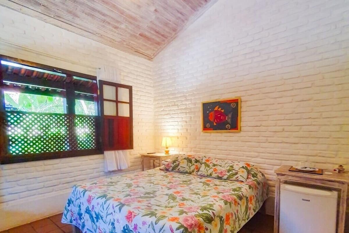 A bedroom with a bed, a nightstand, a lamp, a small refrigerator, and a window with a view of greenery.
