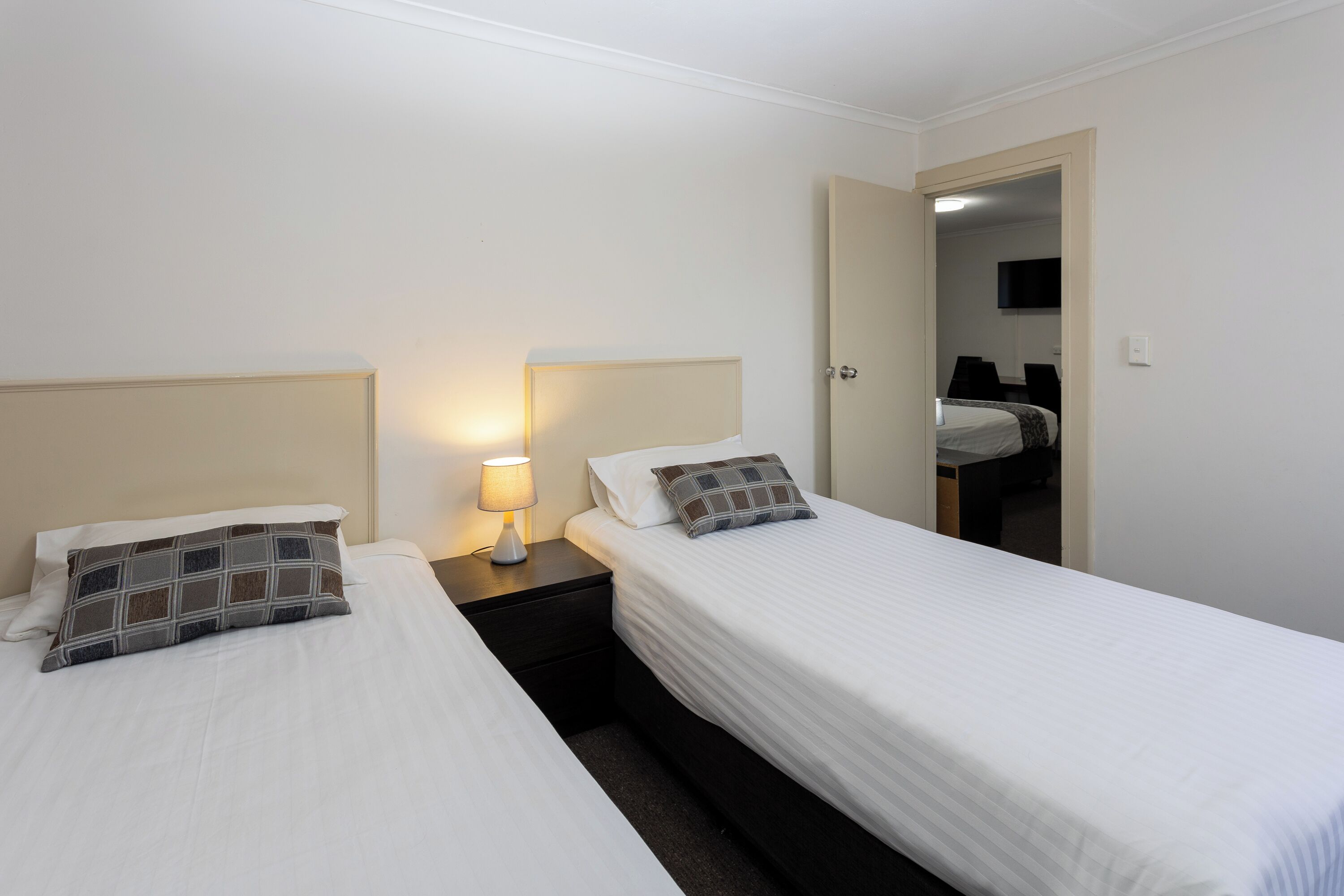 Habitación de hotel con dos camas individuales, una mesita de noche con lámpara, y una puerta que da al baño.