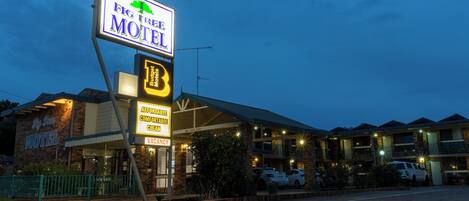 A motel with a large sign, a covered parking area, and multiple buildings in the background.