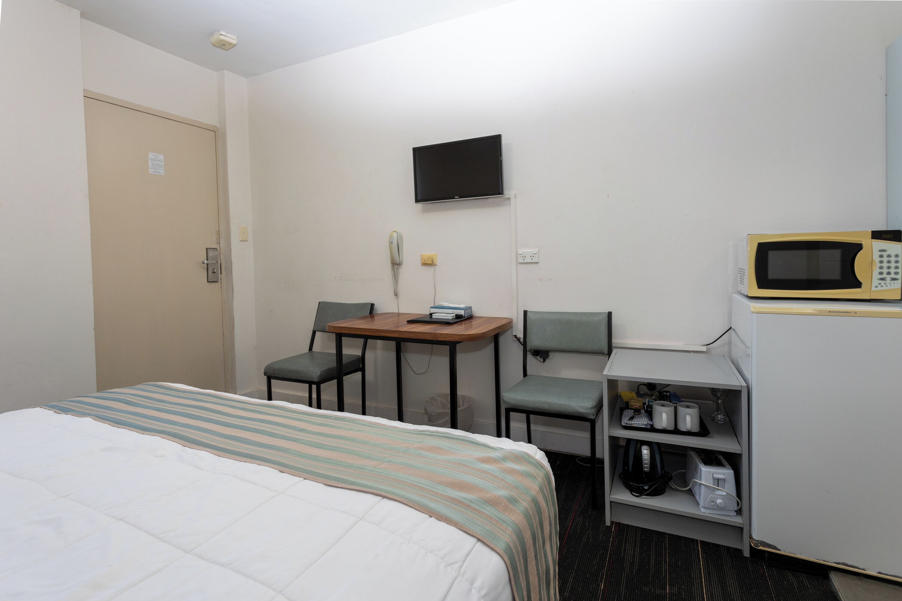 A hotel room with a bed, a desk with a chair, a television, a microwave, and a shelf with various items.