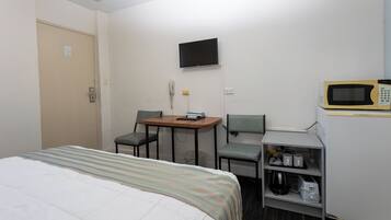 A hotel room with a bed, a desk with a chair, a television, a microwave, and a shelf with various items.
