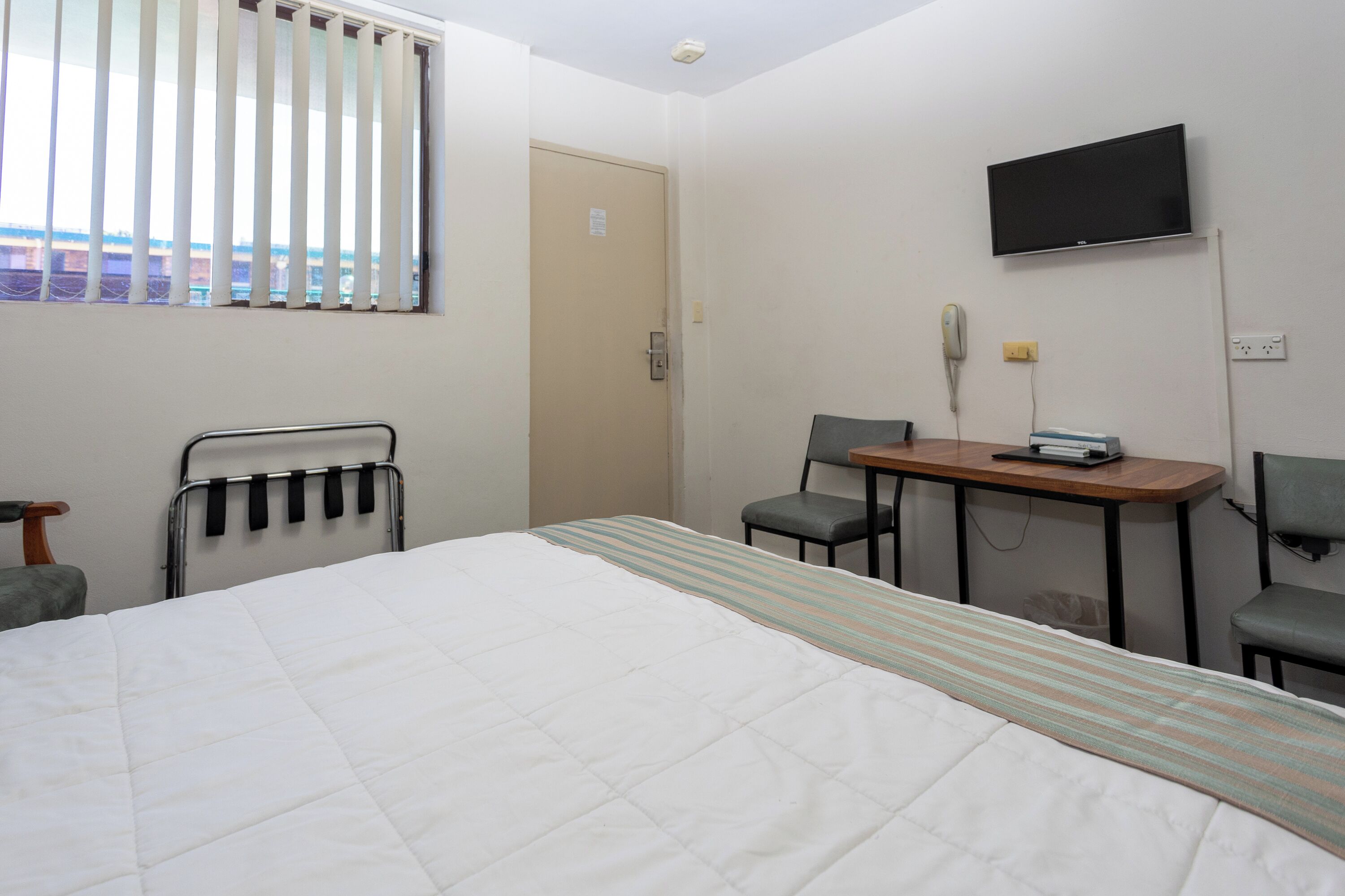 A hotel room with a bed, a desk, two chairs, a television, and a window with blinds.