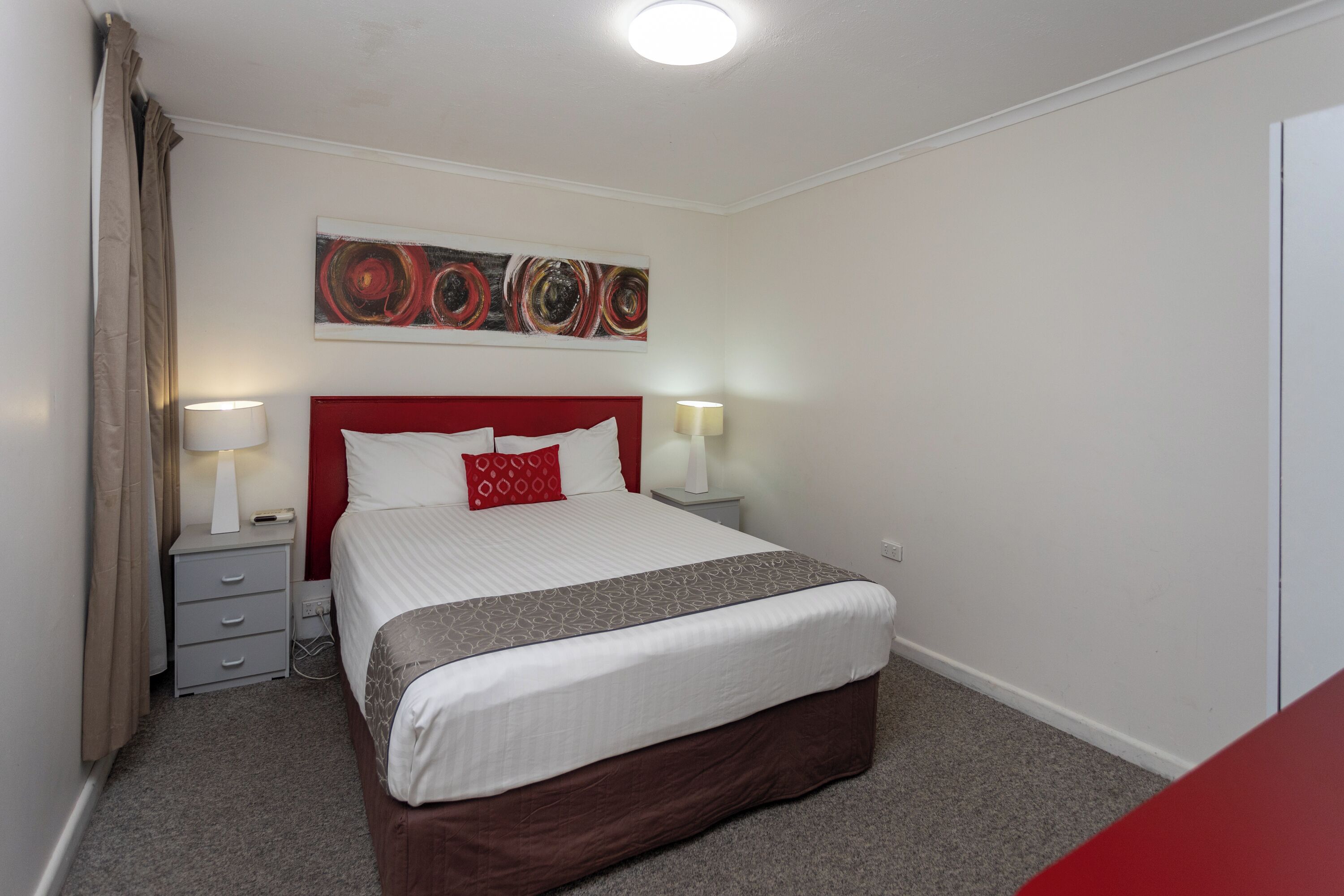 Een netjes ingerichte hotelkamer met een groot bed, twee nachtkastjes met lampen, een rood hoofdbord en een modern abstract schilderij aan de muur.