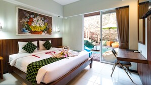 A hotel room with a bed, a desk, a chair, a painting, and a view of an outdoor pool area. - Hadi Poetra Hotel (Kuta)