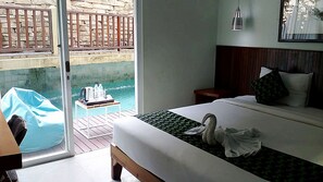A hotel room with a bed, a wooden headboard, a nightstand, a lamp, and a view of a pool through a glass door. - Hadi Poetra Hotel (Kuta)