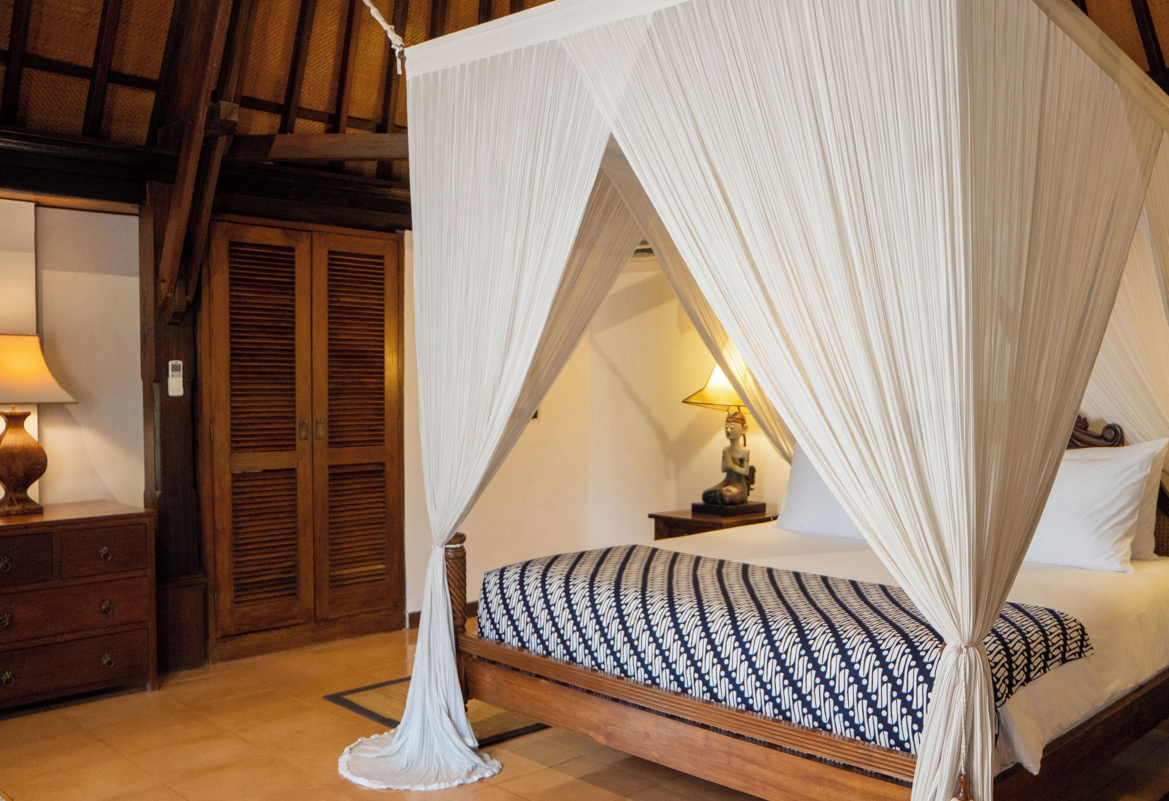 A bedroom with a canopy bed, wooden wardrobe, bedside table with lamp, and a nightstand.