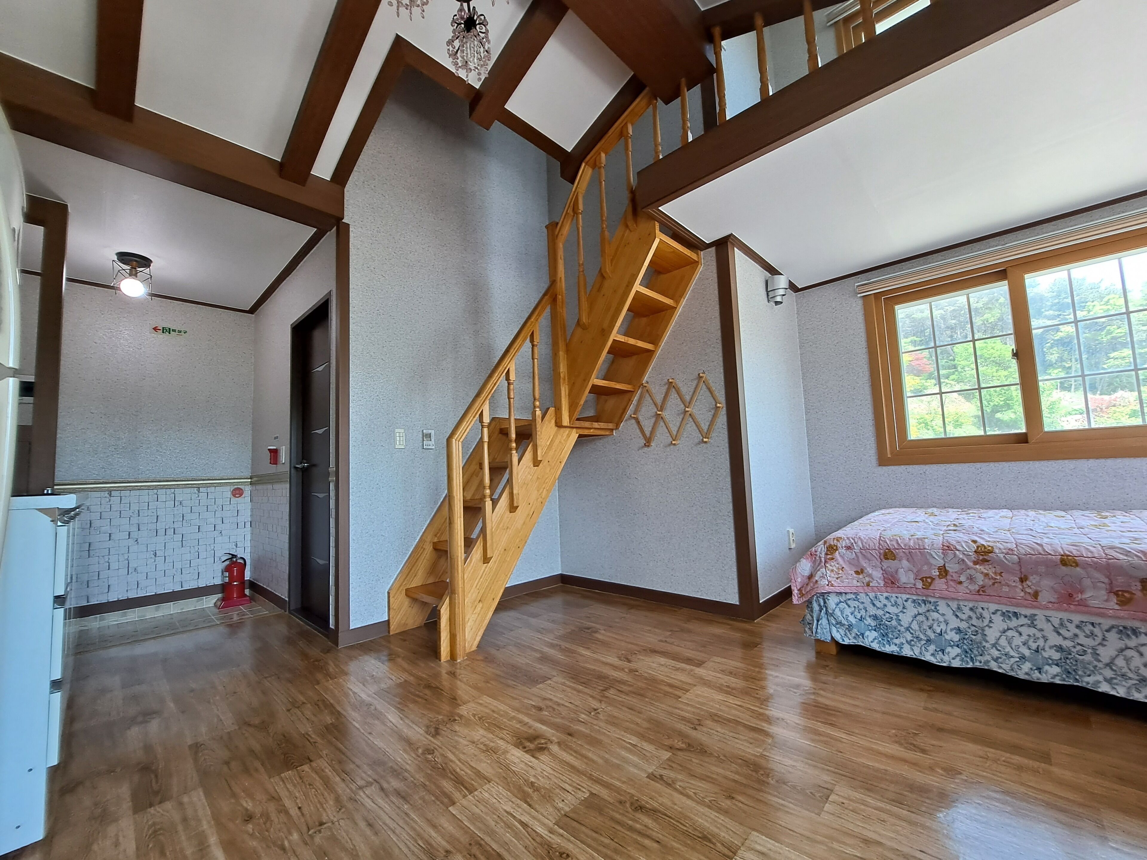 A spacious room with a wooden staircase, a bed with floral bedding, and a large window with a view of greenery.
