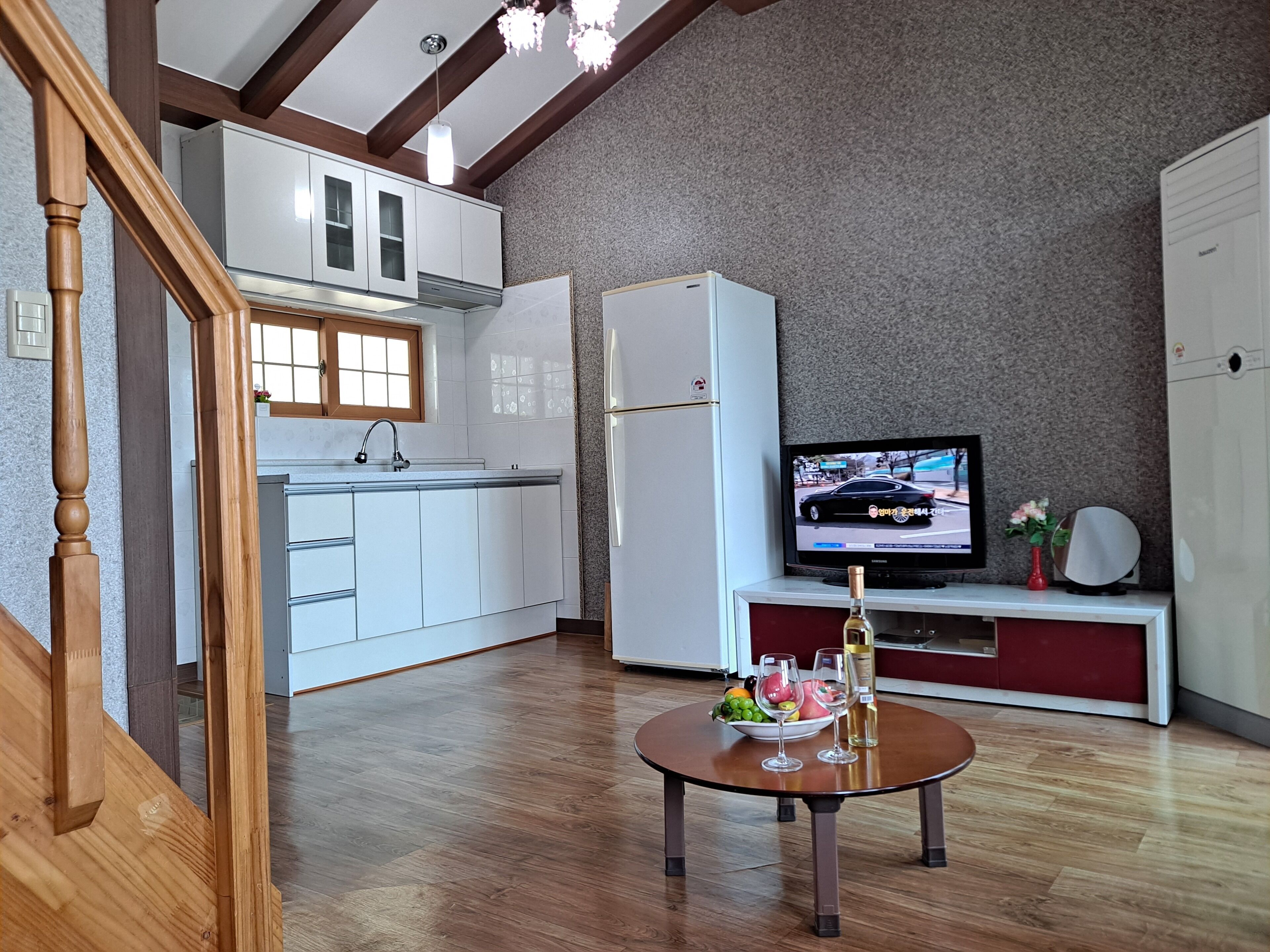 Una cocina moderna con armarios blancos, horno empotrado y refrigerador. Un televisor está montado en la pared, y en primer plano se encuentra una pequeña mesa con un bol de fruta y copas de vino.