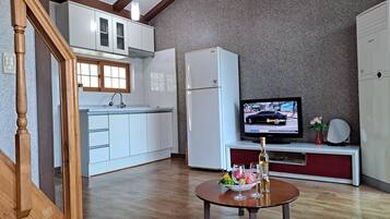 Una cocina moderna con armarios blancos, horno empotrado y refrigerador. Un televisor está montado en la pared, y en primer plano se encuentra una pequeña mesa con un bol de fruta y copas de vino.