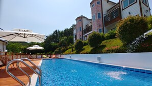 A swimming pool with a wooden deck and metal railings, surrounded by lush greenery and a multi-story building with balconies. - Beljari Pension (Tongyeong)