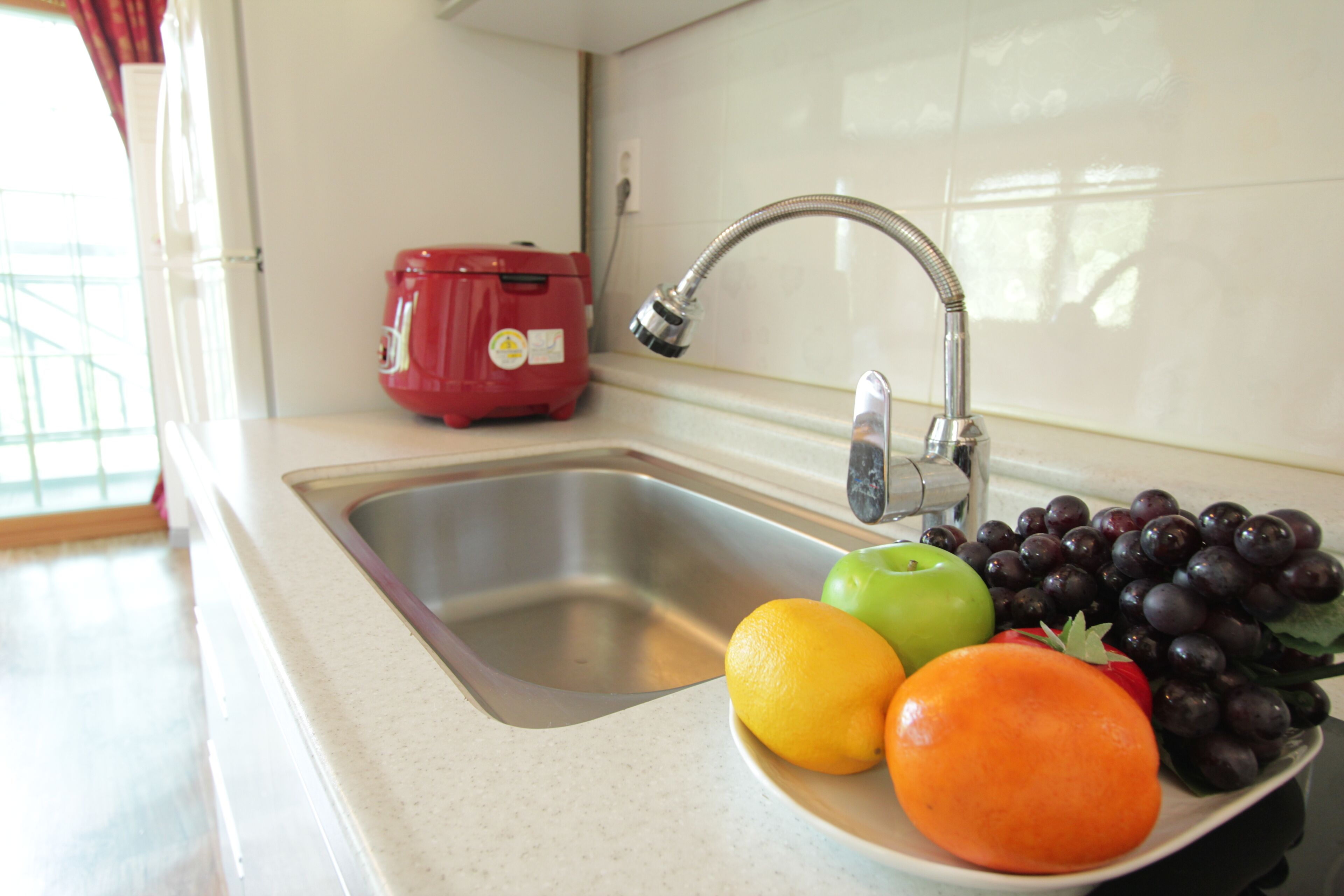 Un lavamanos de cocina con pileta de acero inoxidable, un electrodoméstico rojo y un bol con frutas, incluyendo una manzana, una naranja y uvas.