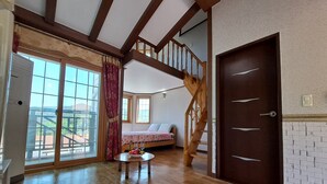 A spacious room with a wooden floor, a bed with a view, a dining table with fruit and wine, and a staircase. - Beljari Pension (Tongyeong)