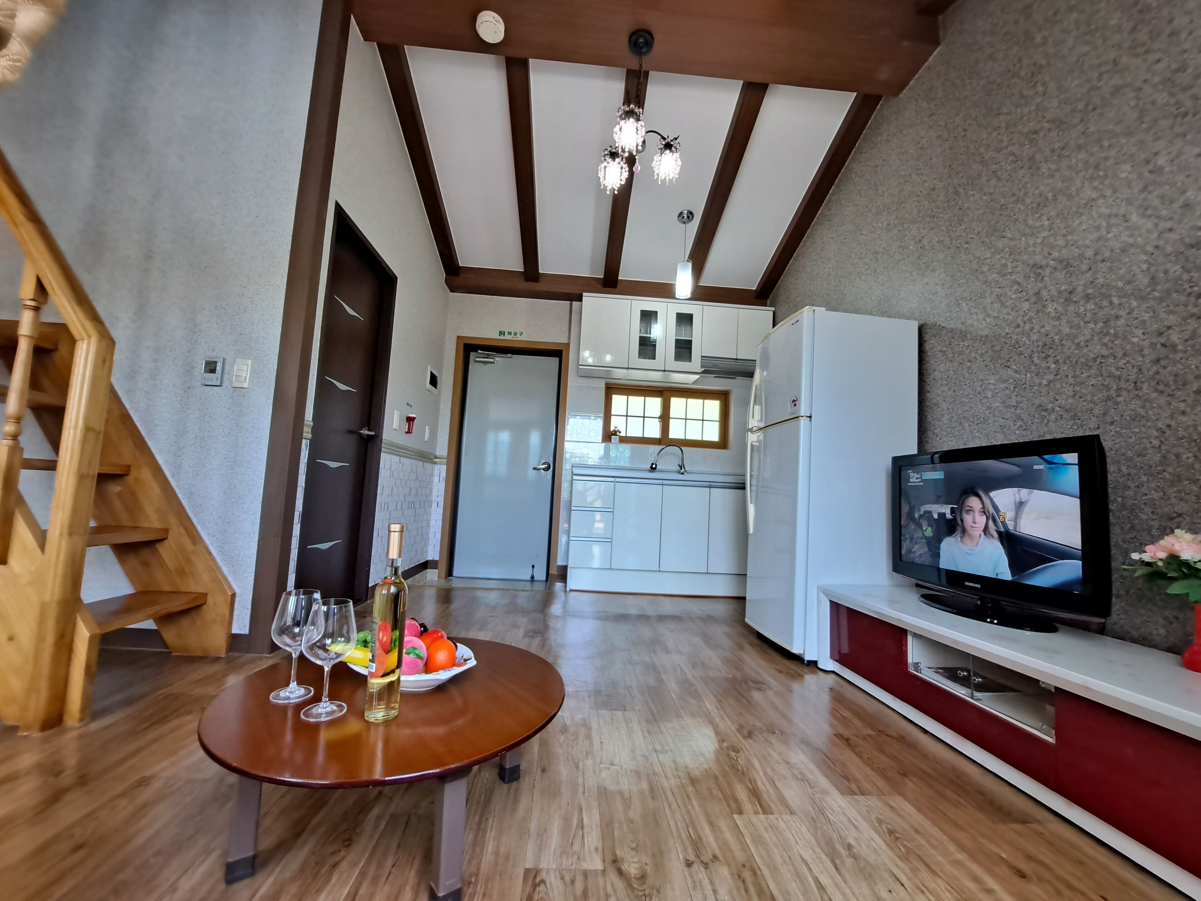 A modern kitchen with a wooden staircase, a dining table with wine glasses and a bottle, and a TV displaying a scene.