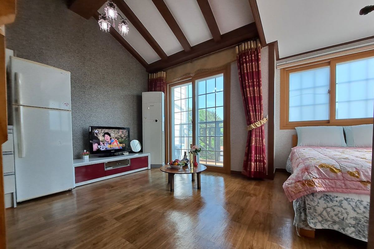 A spacious room with a wooden floor, a bed with a floral bedspread, a television on a red cabinet, a refrigerator, and a large window with curtains.