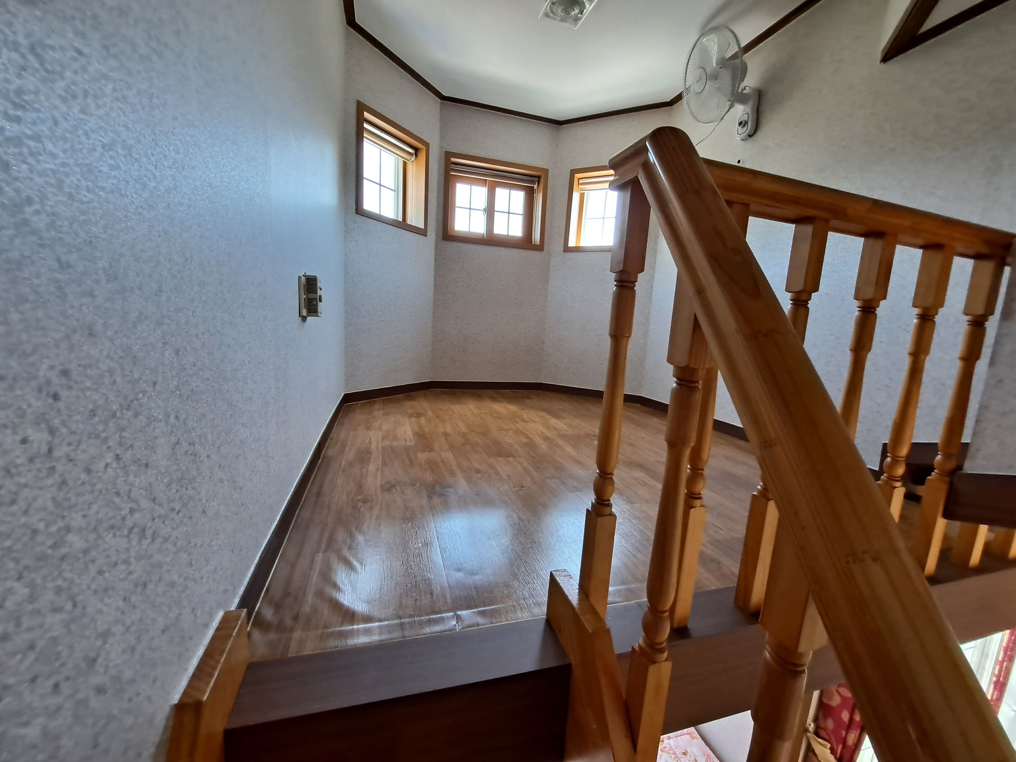 A spacious room with a wooden floor, a ceiling fan, and three windows.