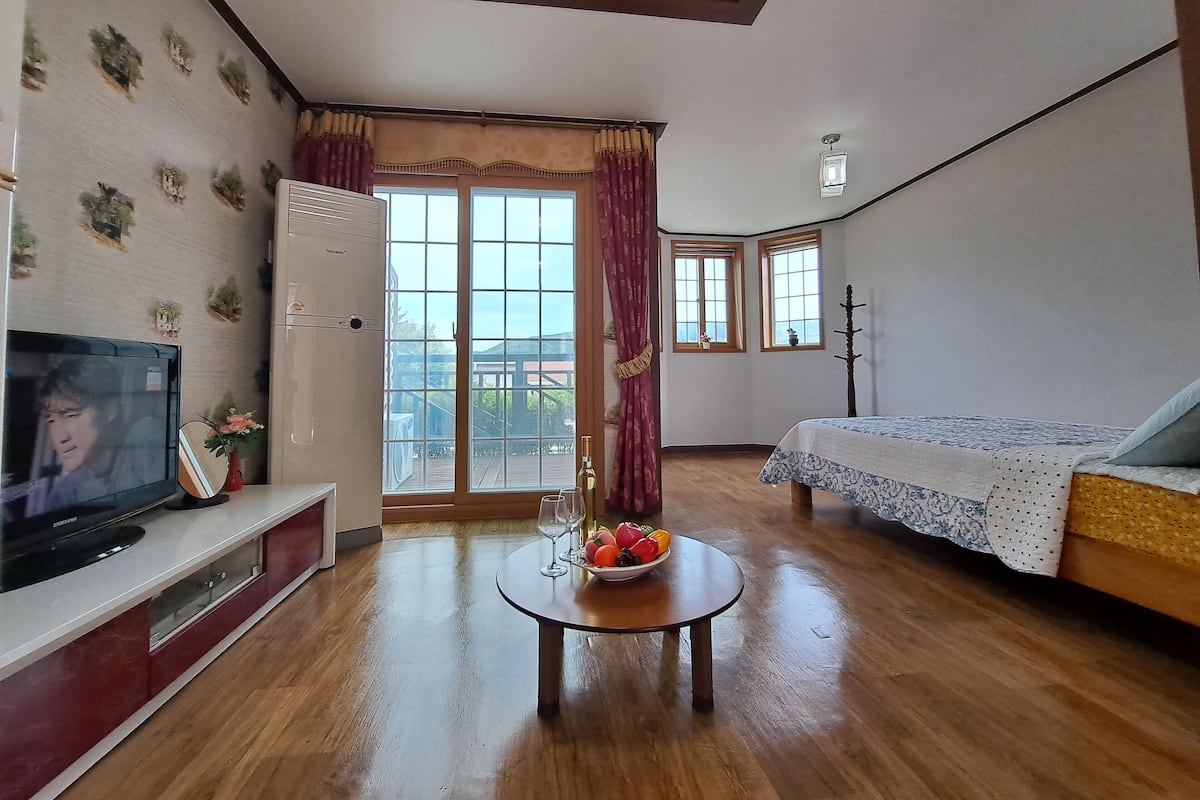 A room with a television, a small table with a bowl of fruit and wine glasses, a sofa, and a bed.