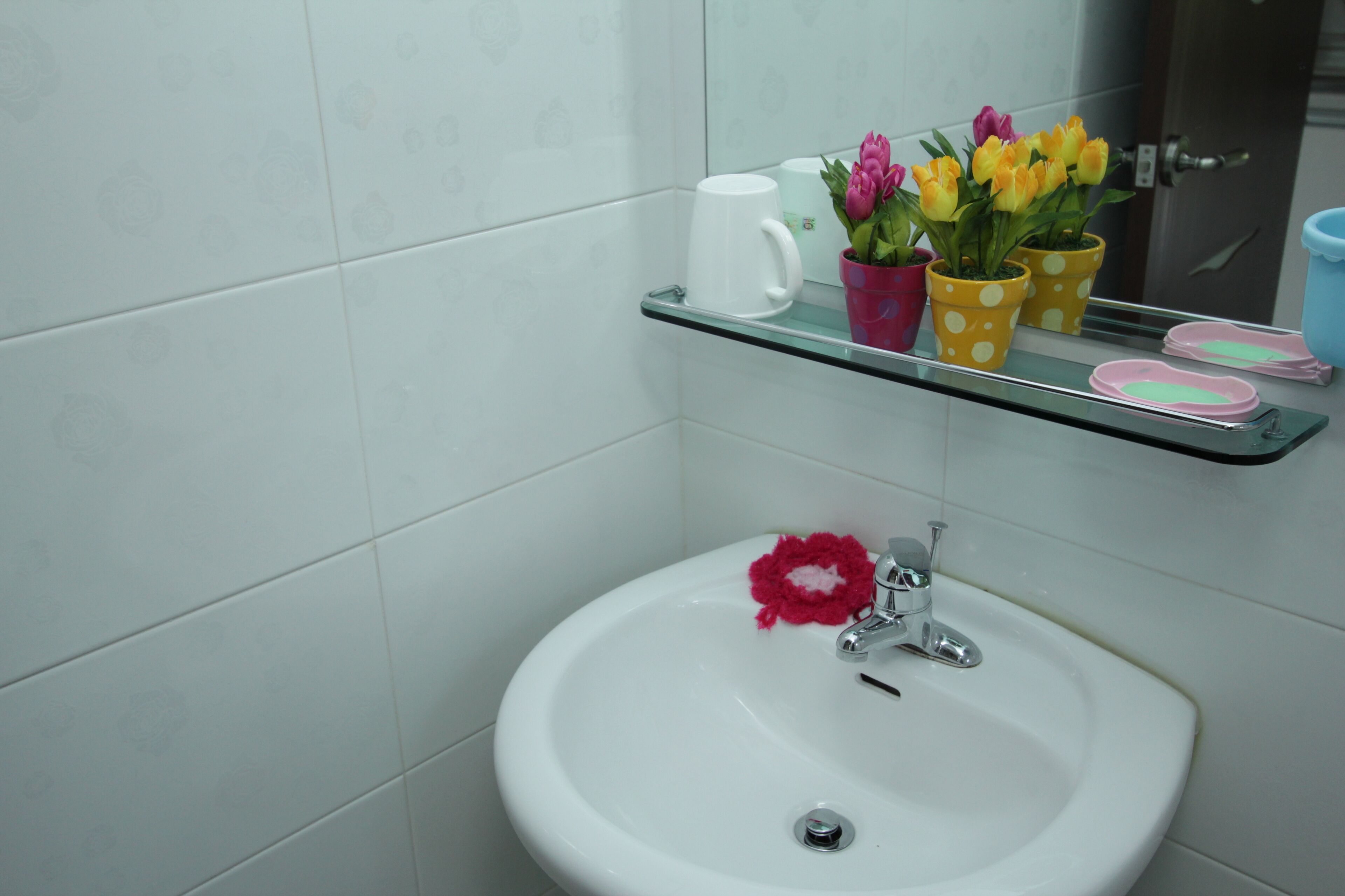 Une salle de bain avec un lavabo blanc, une étagère en verre sur laquelle est posée une tasse blanche, et des tulipes en pot.