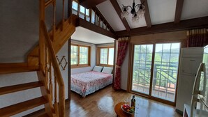 A bedroom with a wooden staircase, a bed with a floral bedspread, a table with fruits and a bottle, and a view of a balcony with a railing. - Beljari Pension (Tongyeong)