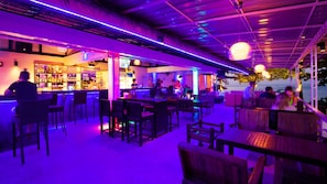 An outdoor bar area with wooden tables and chairs, a bar counter with a bartender, and a view of the sea.