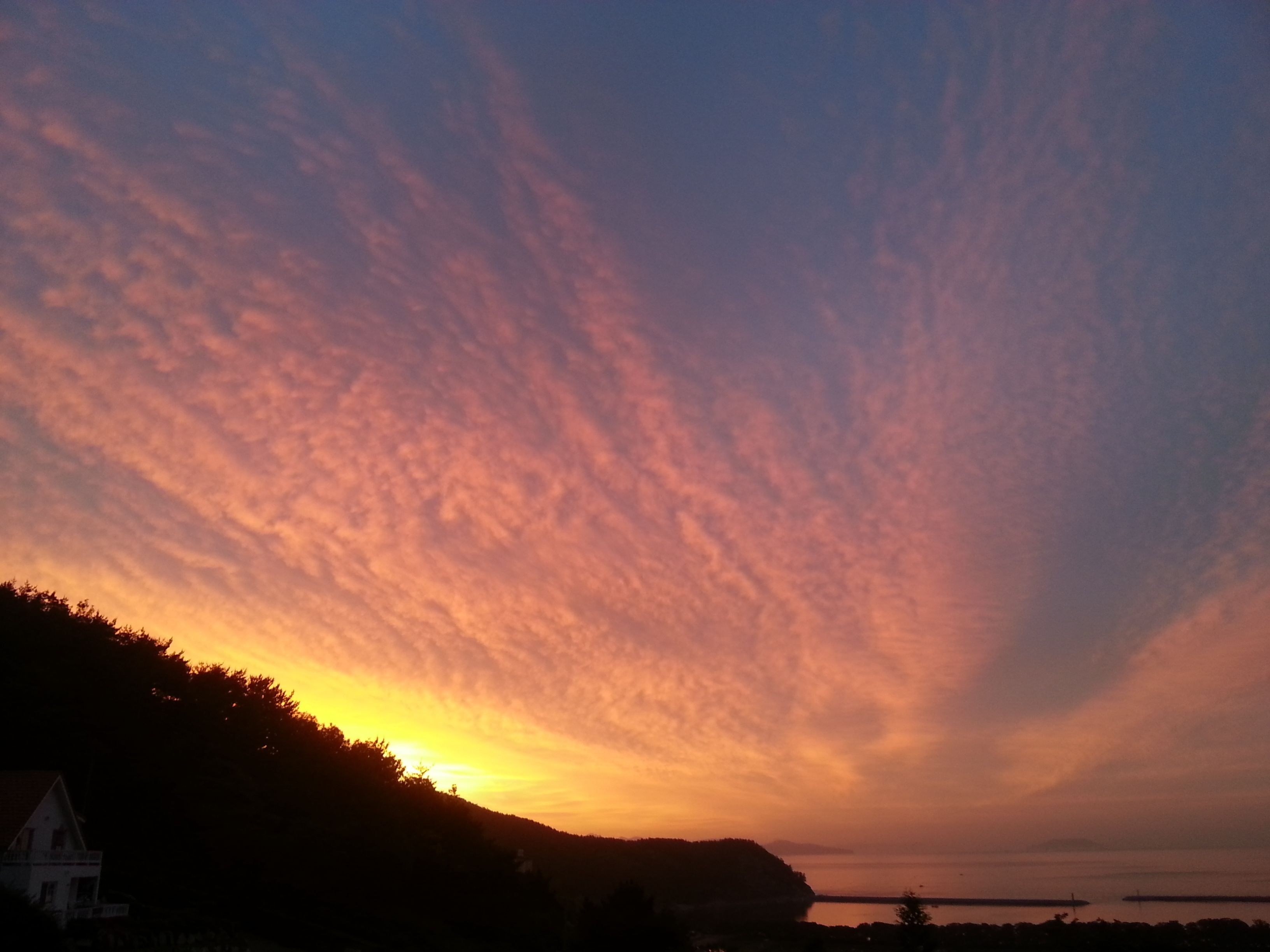 Ein Sonnenuntergang über einer Küstenlandschaft mit der Silhouette eines Hauses und eines Hügels.