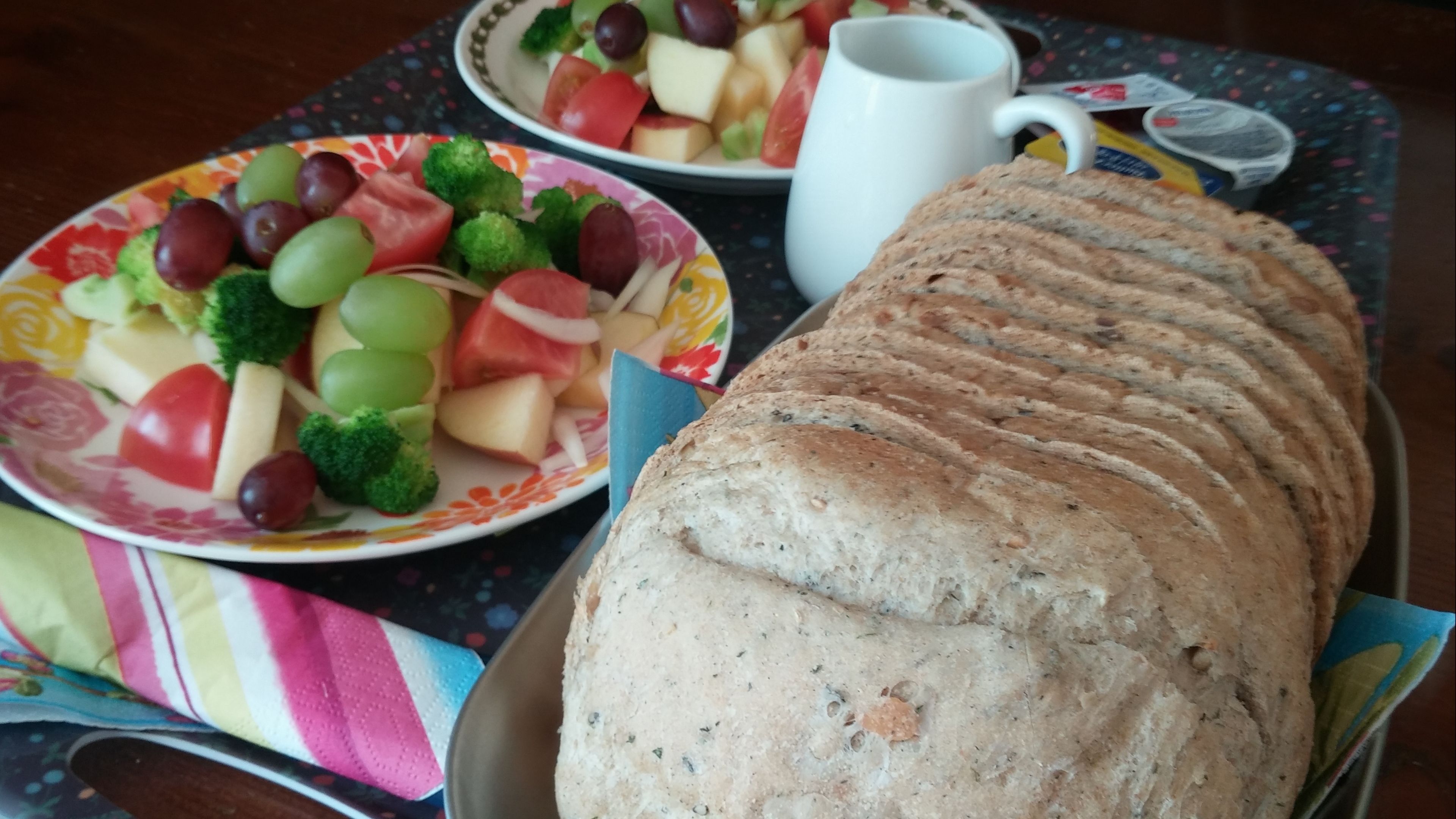 A loaf of bread on a tray, a plate with fruits and vegetables, and a white mug on a table.