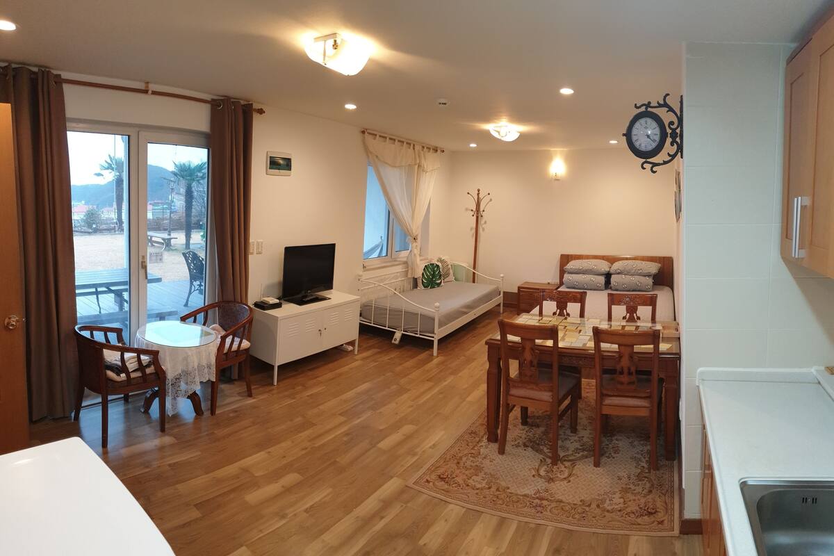 A room with a bed, a TV, a dining table with chairs, a small kitchen area, and a view through a sliding door.