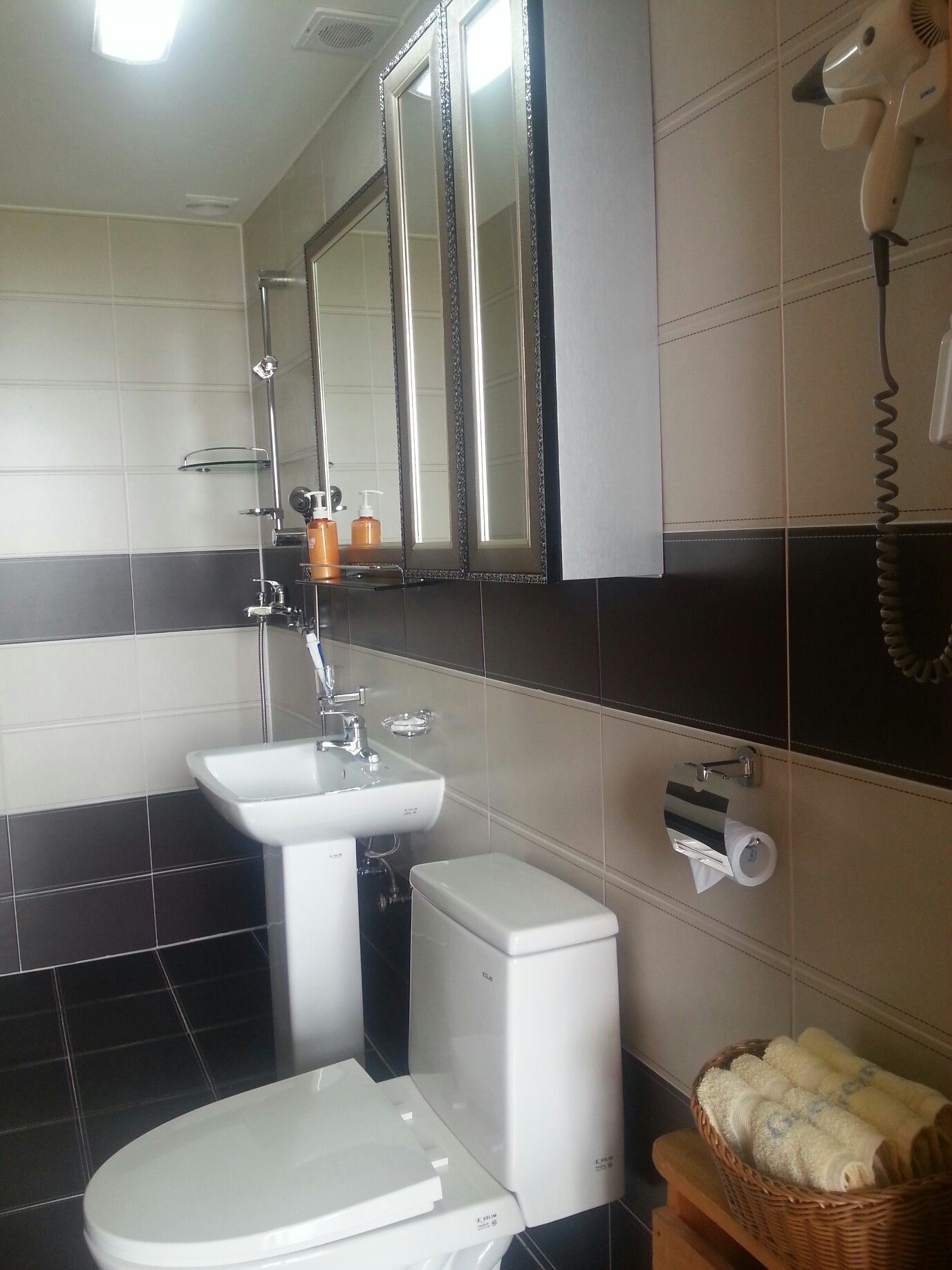 A bathroom with a toilet, sink, and mirror. There is a hairdryer on the wall and a basket of towels on a wooden stool.