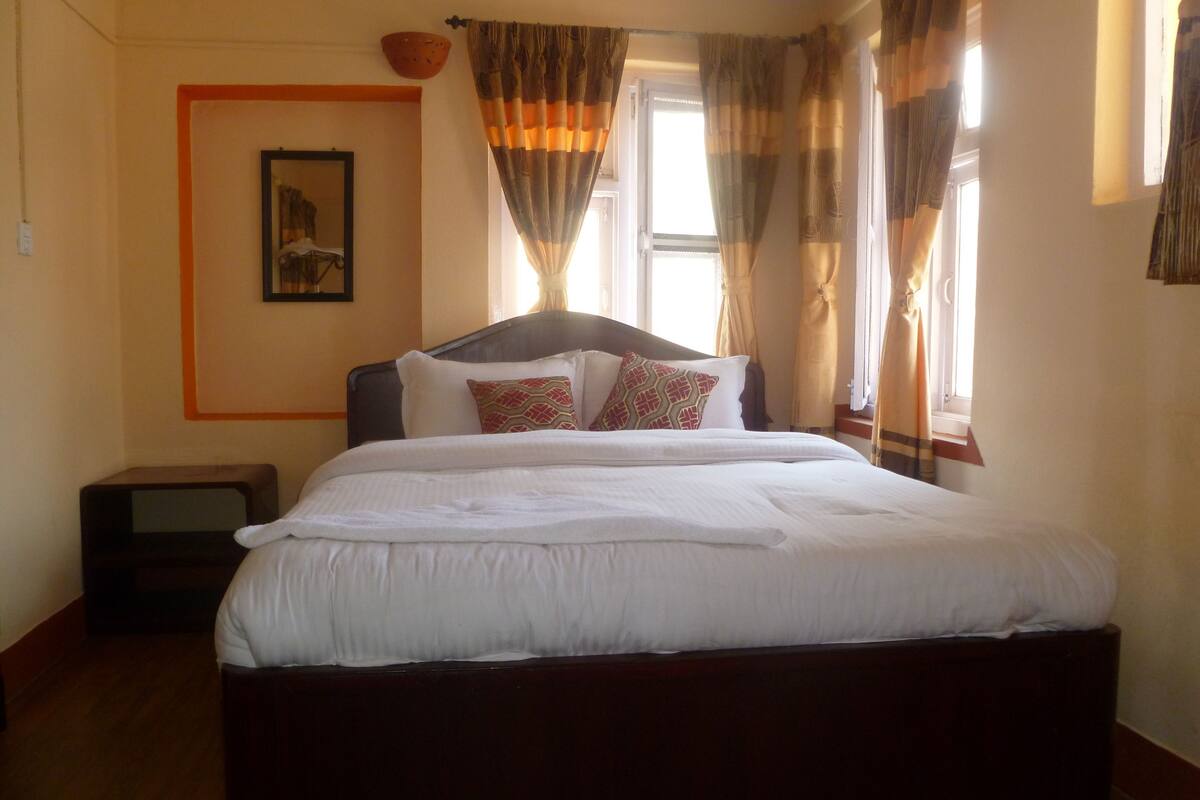A neatly made bed with white linens and patterned pillows, a wooden headboard, and a window with curtains.