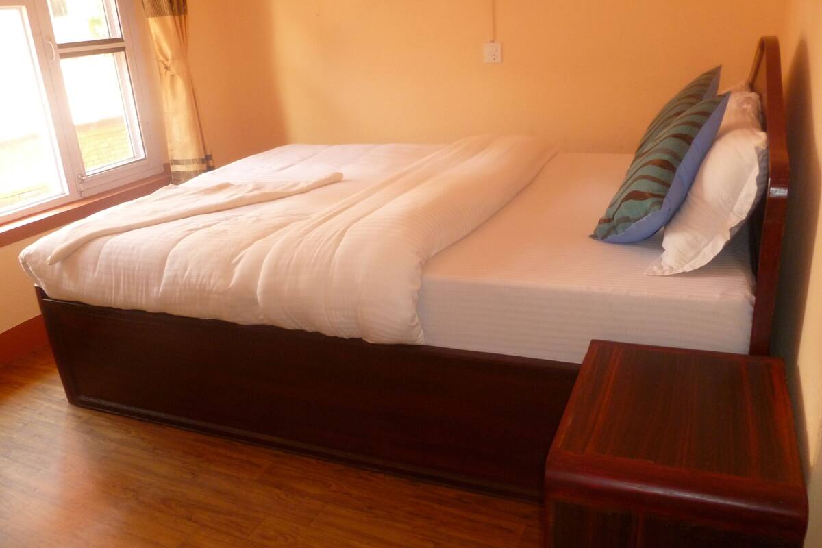 A neatly made bed with a wooden headboard in a room with wooden flooring and a window with curtains.