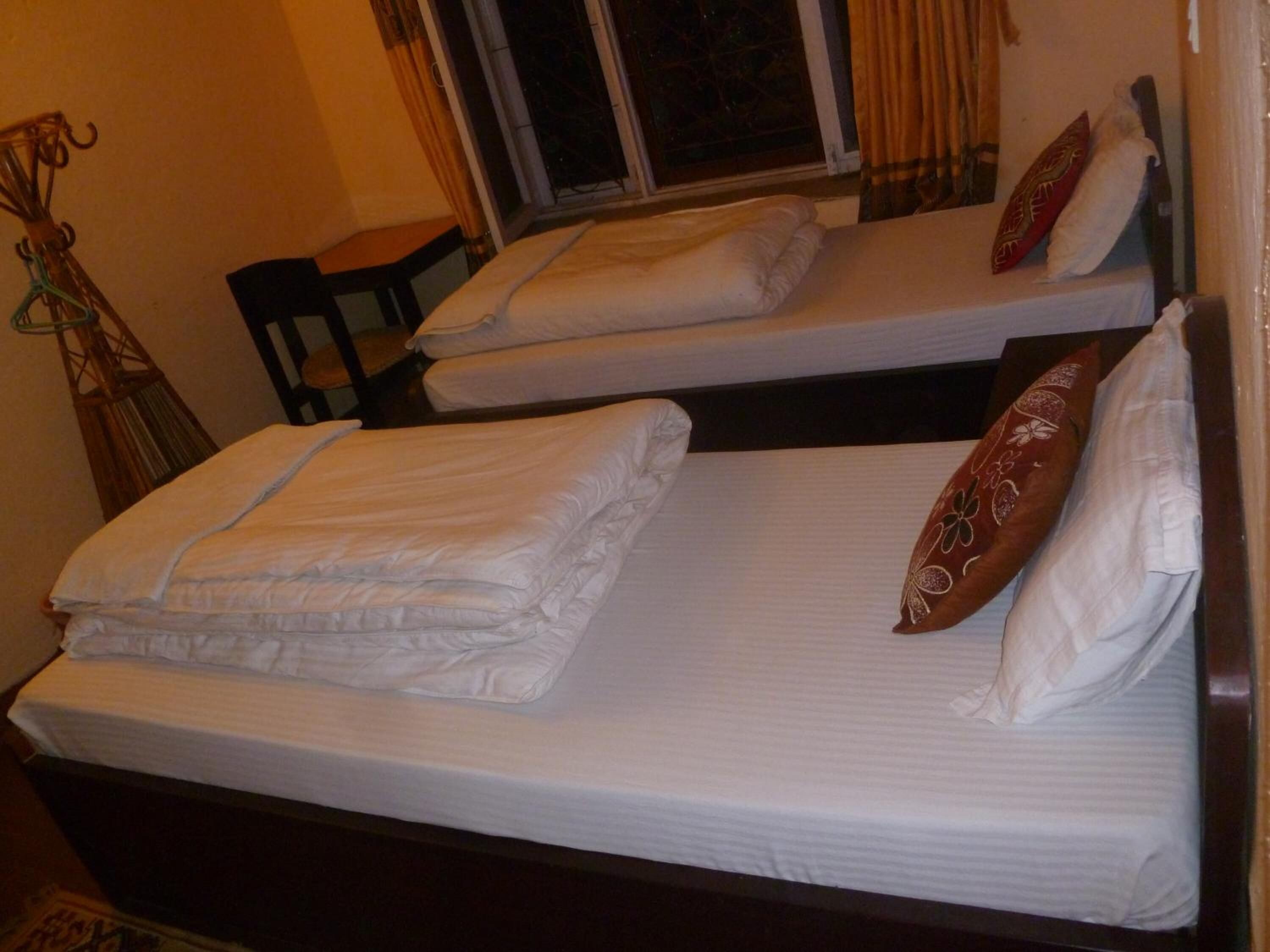 A room with two single beds, each with white linens and pillows. There is a window with curtains, a chair, and a small table.