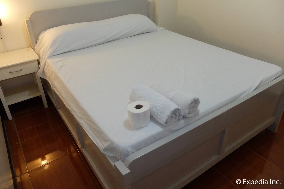 Una cama bien tendida con sábanas blancas y dos toallas enrolladas, una mesita de noche blanca con cajón, y una mesita auxiliar fijada a la pared.