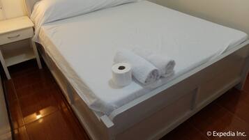 A neatly made bed with white linens and two rolled towels, a white bedside table with a drawer, and a wall-mounted nightstand.