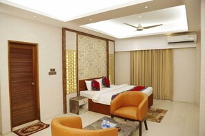 A hotel room with a bed, two chairs, a small table with bottles, a ceiling fan, and an air conditioning unit. - Sea Shell Residence (Dhaka)