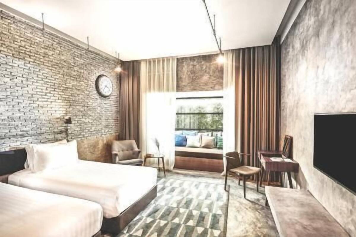 A hotel room with a brick wall, a bed, a sofa, a TV, and a window with curtains.