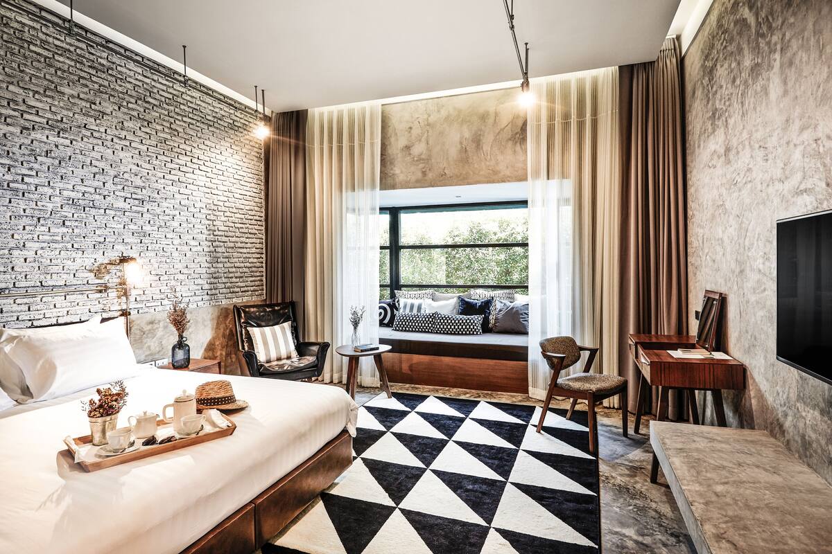 A modern hotel room with a brick wall, a large bed, a black and white checkered floor, a seating area with a view, and a TV on the wall.