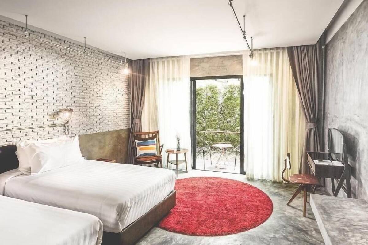 A hotel room with a brick wall, a large bed, a red rug, a balcony with a table and chairs, and a view of greenery outside.