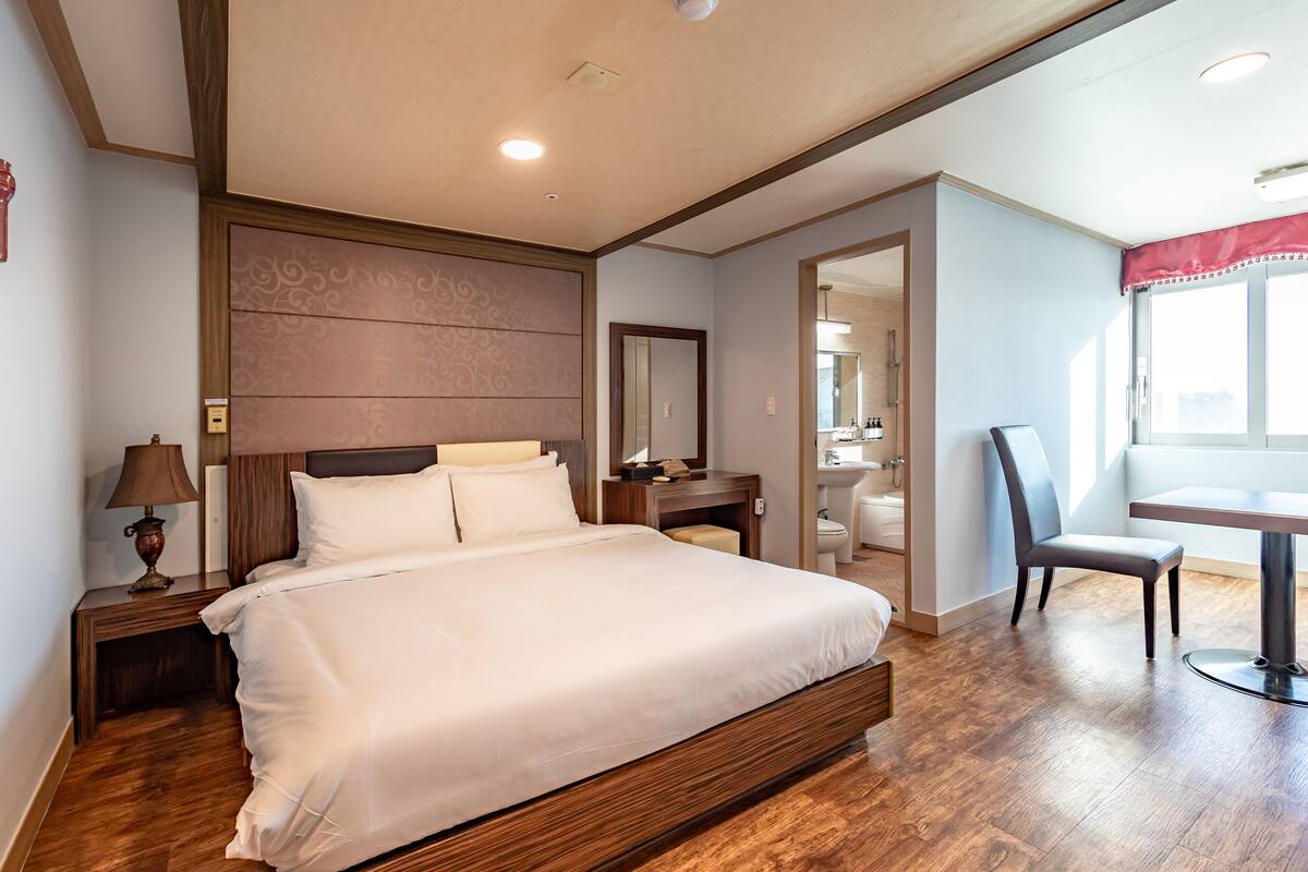 Una habitación de hotel con una cama grande, una mesita de noche con lámpara, un escritorio con silla y una ventana con vistas a la ciudad.