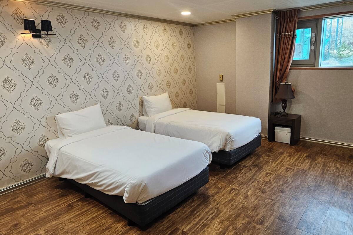 Habitación de hotel con dos camas, una mesita de noche con lámpara, ventana con cortinas y papel tapiz estampado.