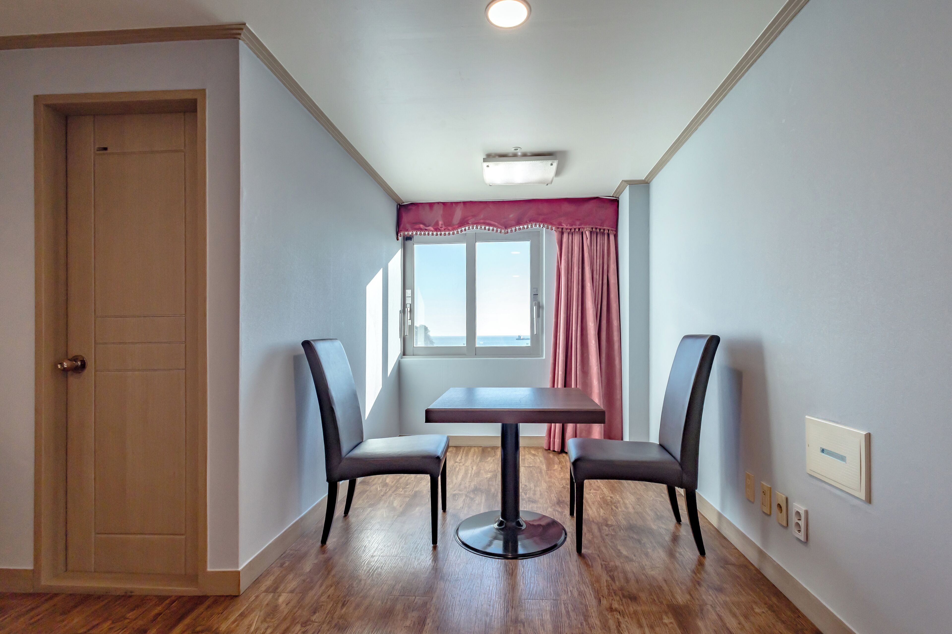 A room with a wooden door, a table with two chairs, a window with a view, and a ceiling light.