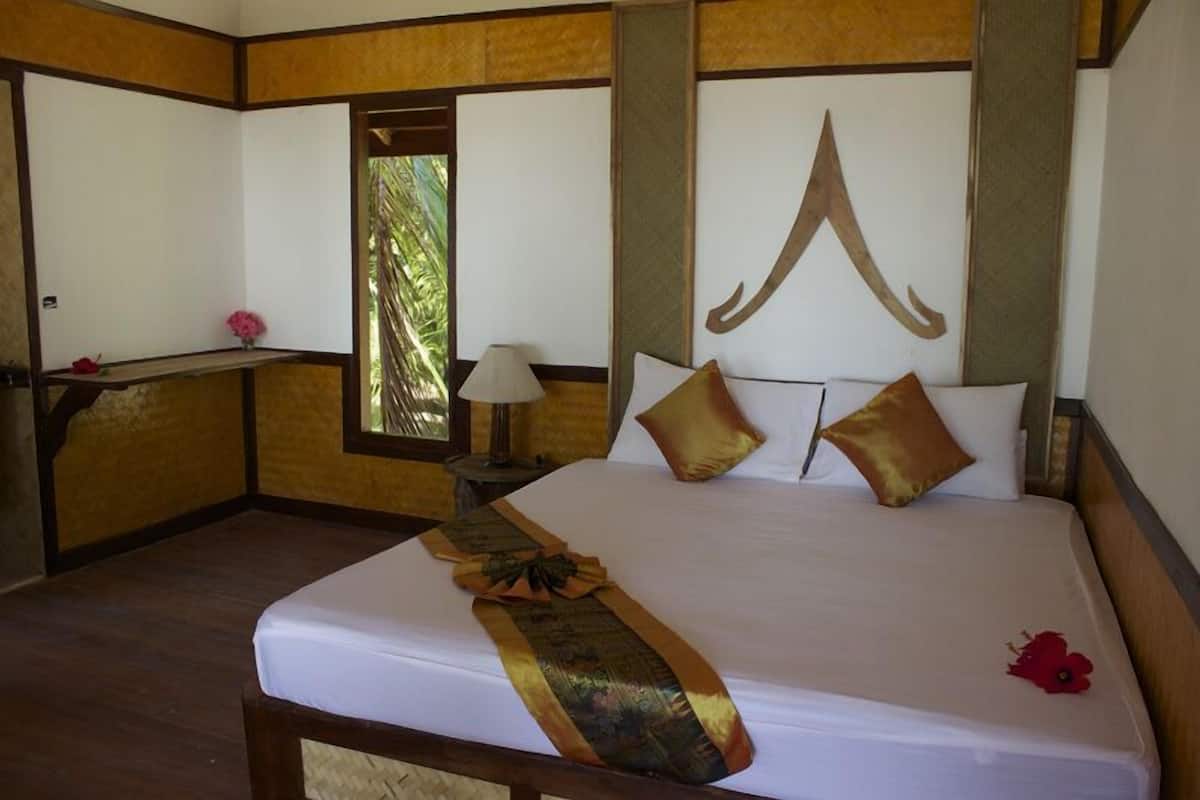 A hotel room with a large bed, a nightstand, a lamp, and a window with a view of palm trees.