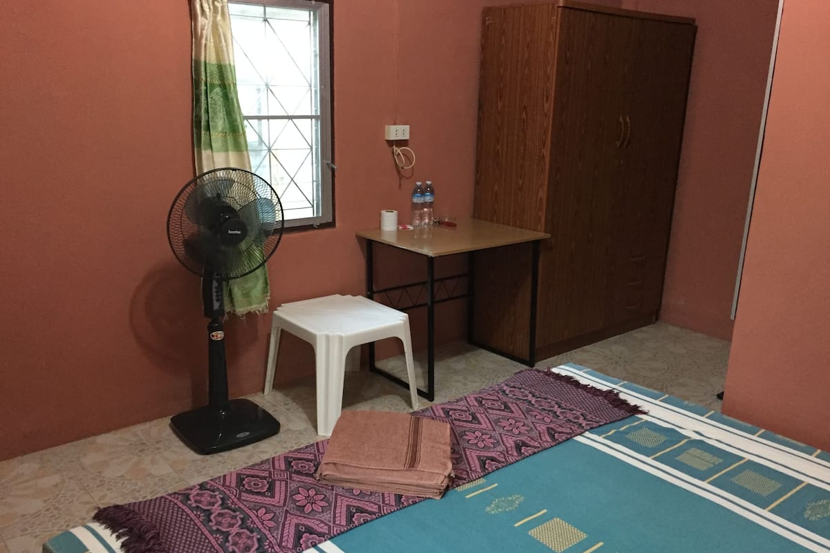 Una habitación pequeña con cama, ventilador, una mesita, armario y ventana con cortinas.