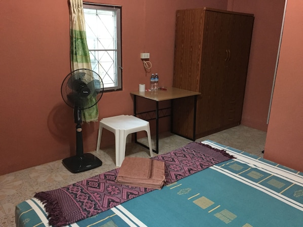 Una habitación pequeña con cama, ventilador, una mesita, armario y ventana con cortinas.