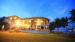 Un grand bâtiment circulaire doté d’un espace extérieur couvert, entouré de palmiers et bordé par une plage.