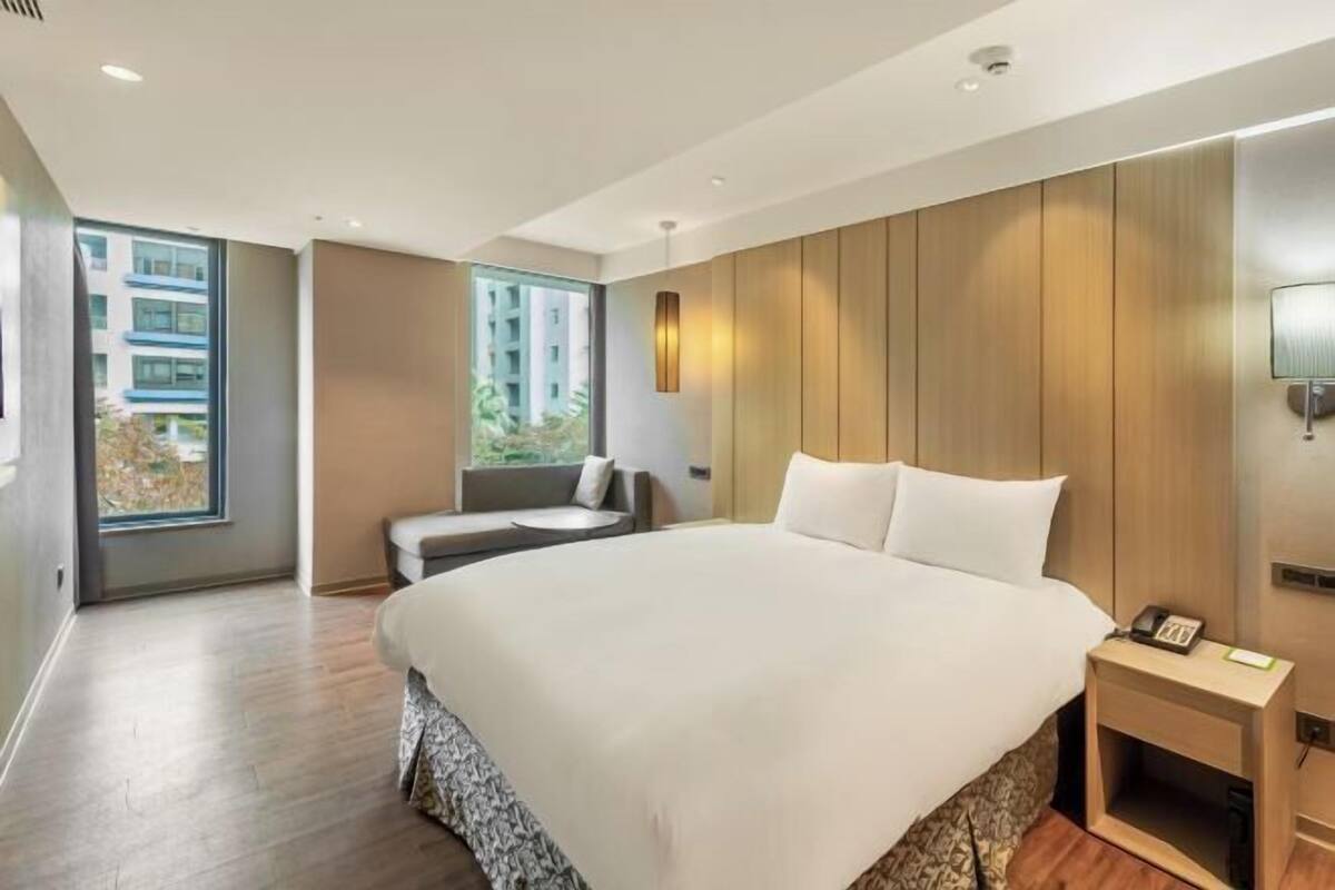 A modern hotel room with a large bed, a bedside table, a lamp, and a view of the outside.