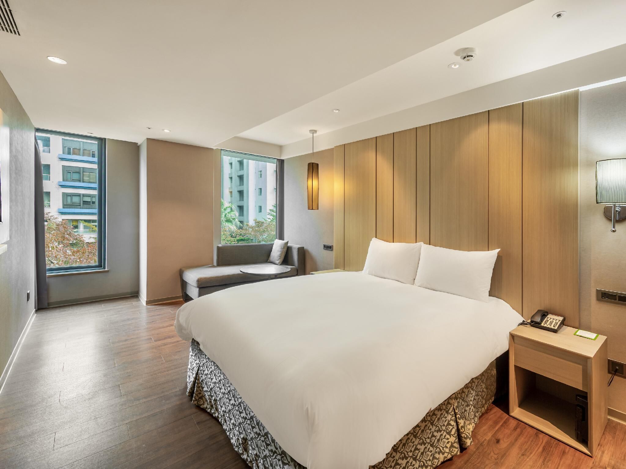 A modern hotel room with a large bed, a seating area, a bedside table with a phone, and a window with a view of buildings.