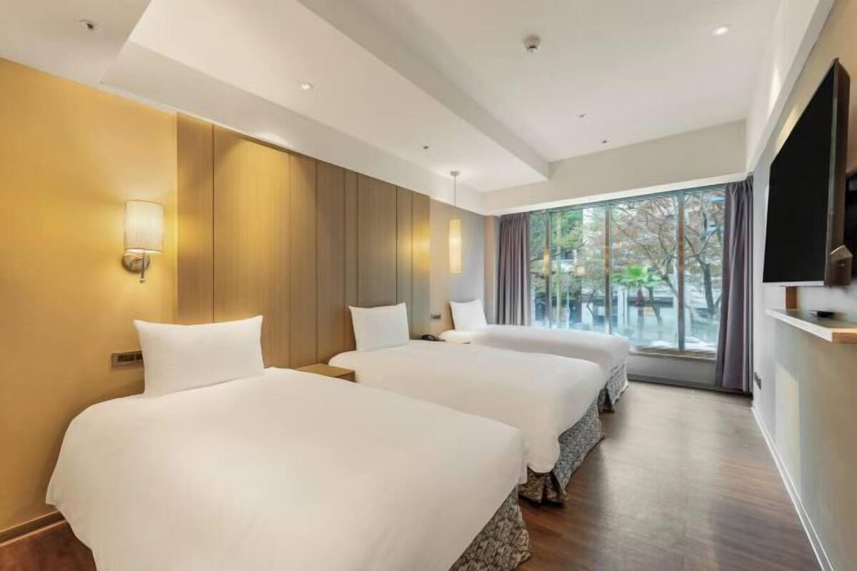 A hotel room with two beds, a large window with a view, and a flat-screen TV mounted on the wall.