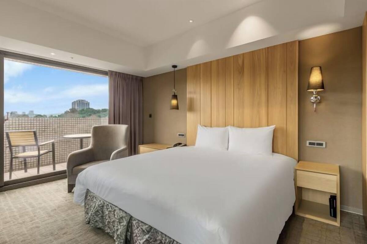 A hotel room with a large bed, two bedside tables, a chair, a small table, and a view of the city through large windows.