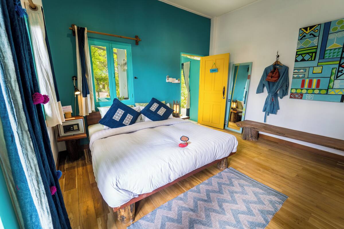 A bedroom with a bed, a wooden bench, a blue and white patterned rug, and a colorful abstract painting on the wall.