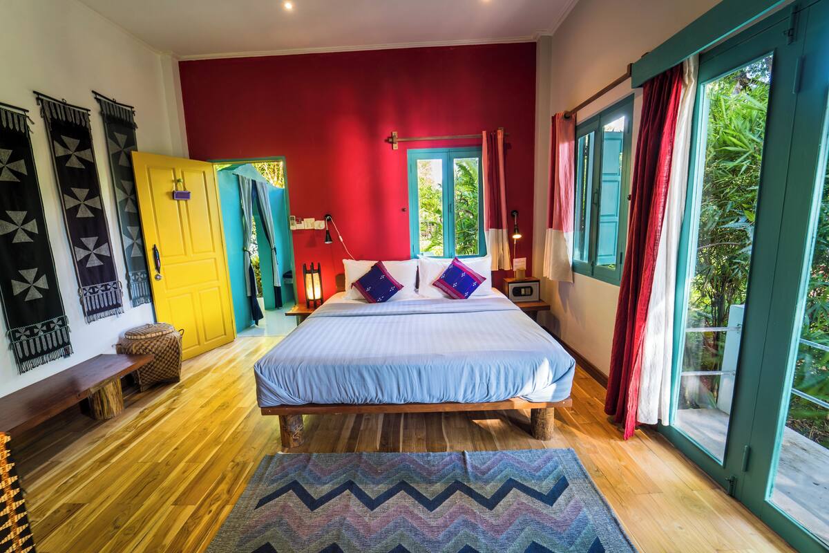 A bedroom with a bed, wooden floor, a bench, a window with curtains, and a door.
