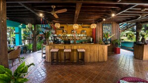Uma área de bar com balcão de madeira, três banquetas, luminárias pendentes em formato de esfera e um ventilador de teto.